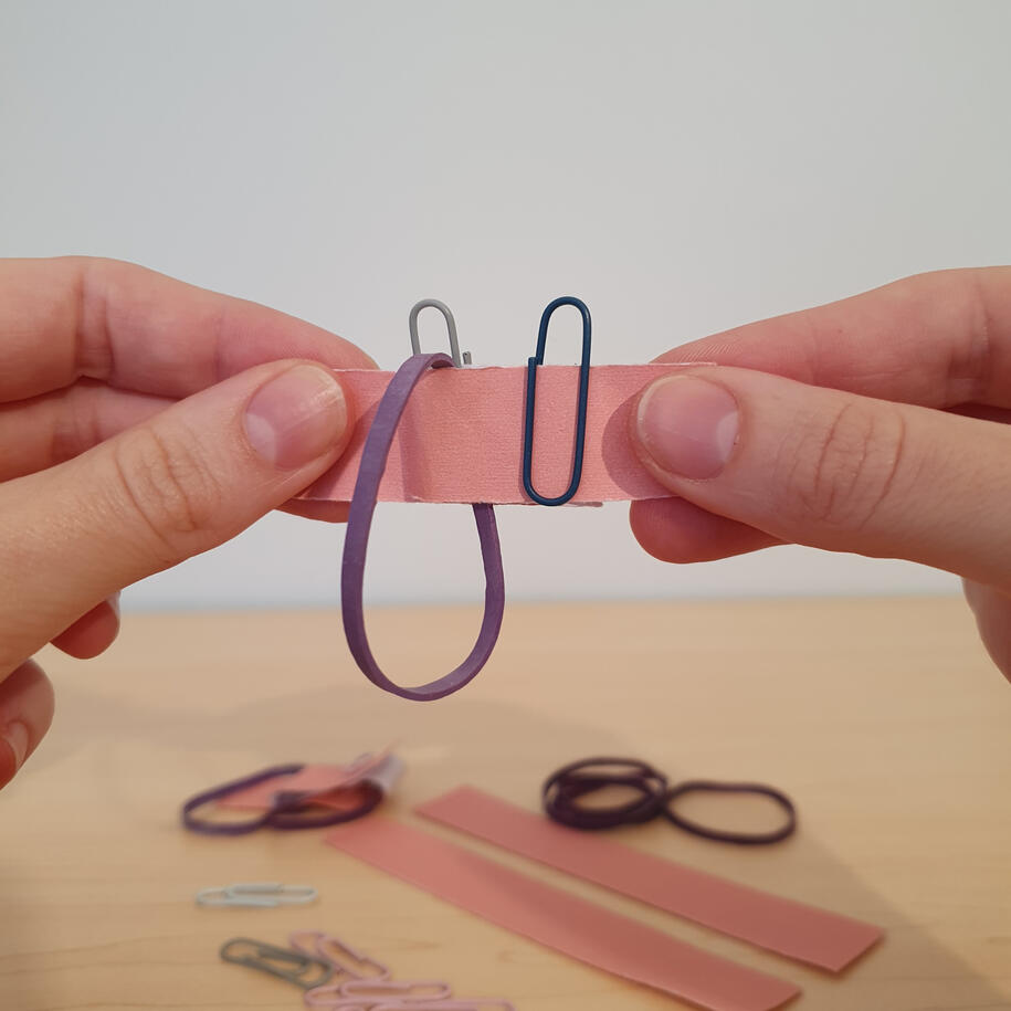 Paperclip magic trick for primary students investigation Hands linking paperclips and a rubber band on a folded paper strip, a topology magic trick showing how loops link when pulled taut